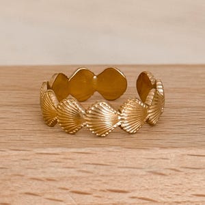 May include: A gold-colored ring featuring a seashell design. The ring is made of a shiny metal and has a series of detailed seashell shapes around the band. The ring is displayed on a light-colored wooden surface.