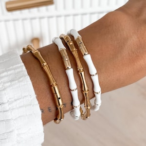 May include: Four gold and white bamboo-style bracelets are stacked on a wrist. The bracelets have a gold clasp and a white enamel center. The background is a light wood surface and a white shirt.