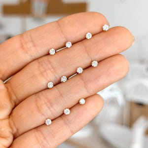 May include: A collection of small, round, clear stud earrings with silver-toned settings. Each earring features a single faceted stone that catches the light, arranged in a series across the fingers to display their size and sparkling appearance.