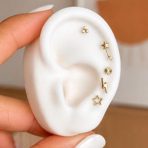 May include: A white ear-shaped display model featuring five gold-toned stud earrings. The set includes a three-stone cluster, a curved bar with a star and crystal, a sunburst disc, a lightning bolt, and an open-frame star stud earring design.
