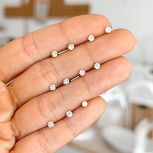 Op de afbeelding: Een verzameling kleine, goudkleurige oorstekers en barbell oorbellen, elk met een enkele, heldere, ronde edelsteen. De oorbellen worden gepresenteerd tegen een lichte achtergrond, die hun delicate ontwerp en glans benadrukt. De afbeelding toont de minimalistische stijl van de sieraden.