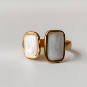 May include: A gold-toned ring featuring two rectangular, side-by-side settings. One setting is filled with white material, and the other with a light gray material. The ring is set against a white background.
