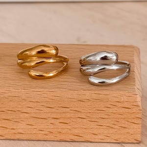May include: Two abstract rings, one gold-toned and one silver-toned, are displayed on a wooden block. The rings have a fluid, organic design, with a wrap-around style. The gold ring is on the left, and the silver ring is on the right.