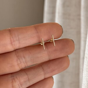 May include: A pair of small gold stud earrings, each featuring a thin, vertical bar design. The earrings are polished and metallic, with a simple straight shape that sits horizontally across the fingers in this view.