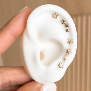 以下が含まれることがあります： 白い耳のモデルに展示された、金色の星型のイヤリングのコレクション。 イヤリングは、無地の星、テクスチャード加工の星、小さな宝石が付いた湾曲したイヤリングなど、デザインが異なります。 背景はニュートラルベージュです。