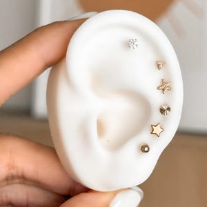 May include: A white ear-shaped display model featuring six gold-toned stud earrings arranged in a vertical line. The set includes a clear crystal stud, a small triangle, a textured starfish, a sunburst disc, a smooth star, and a round ball stud.