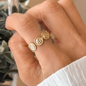 May include: A gold-colored ring featuring a series of circular medallions. Each medallion has a detailed design, and the ring is worn on a finger. The ring's design is visible against the backdrop of a person's skin and a white shirt.