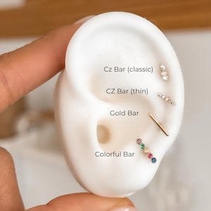 以下が含まれることがあります： さまざまな種類のイヤリングが付いた白い耳のモデルのクローズアップ。イヤリングには、ゴールドバー、CZバー（クラシック）、CZバー（細い）、複数の色の宝石が付いたカラフルバーが含まれます。モデルのテキストは各イヤリングにラベルを付けています。