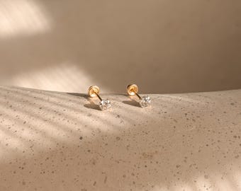 May include: A pair of gold stud earrings, each featuring a single clear, round-cut gemstone set in a four-prong gold setting with a thin gold post and a small, round gold backing, displayed as a matching set of two individual earrings.