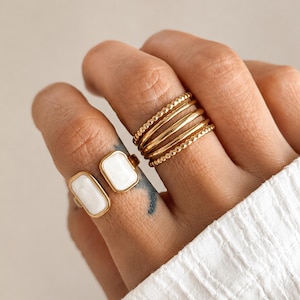 May include: A gold-toned open ring featuring two rectangular white enamel inlays, paired with a stack of five gold-toned rings including two beaded bands and three smooth bands, worn on the fingers of a hand against a neutral background.