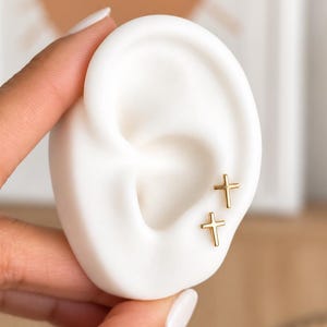 May include: Two small gold-toned cross stud earrings are displayed on a white silicone ear model. The earrings feature a simple, polished metal design with a classic cross shape, positioned vertically on the lobe and cartilage area of the ear.