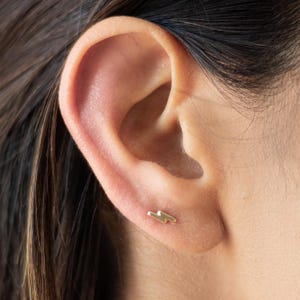 May include: A close-up of a person's ear with a small gold lightning bolt stud earring.