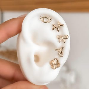 May include: Gold stud earrings in the shapes of an eye, a balloon dog, a bow, a hummingbird, and a flower. The earrings are displayed on a white ear-shaped form.