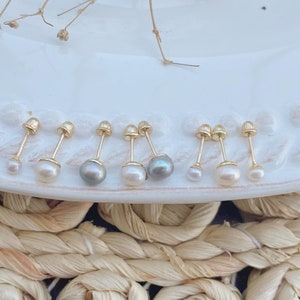 May include: A collection of gold stud earrings with pearl accents. The earrings vary in size and pearl color, including white and gray pearls. The earrings are displayed on a white plate with a woven straw mat in the background.