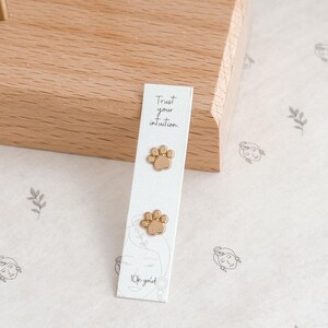 May include: Gold-colored paw print stud earrings displayed on a white card with the text "Trust your intuition" and "10k gold". The earrings are presented on a light wooden surface, suggesting a jewelry display.