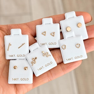 May include: A collection of six pairs of 14kt gold stud earrings displayed on individual white cards. The designs include small circles, triangles, bars, hearts, and snakes. Each white card is clearly labelled with the text 14KT. GOLD.