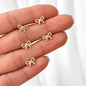 Könnte beinhalten: Vier kleine Ohrstecker aus goldfarbenem Metall, die jeweils wie eine zarte Schleife mit einer glatten, polierten Oberfläche geformt sind. Die Ohrringe haben ein einfaches, symmetrisches Design mit zwei Schlaufen und zwei kurzen Enden.