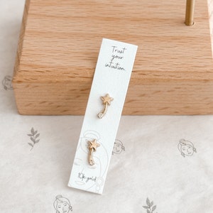 Puede incluir: Un par de pendientes dorados con forma de estrella y un acento curvo y brillante. Los pendientes se muestran en una tarjeta blanca con el texto "Trust your intuition" y "10k gold."