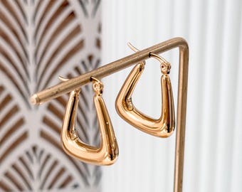 Op de afbeelding: Een paar gouden creolen met een geometrische, driehoekige vorm. De oorbellen worden gepresenteerd op een gouden standaard.