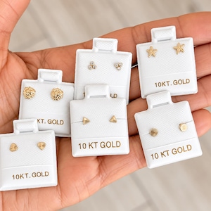May include: Six pairs of 10kt gold stud earrings displayed on white cards. Designs include textured spheres, three-stone clusters, starfish, triangles, fan shapes, and flat discs. Each card is clearly labeled with the text 10KT. GOLD.
