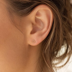 May include: A small, gold-colored stud earring in the shape of a simple, open triangle. The earring is positioned in the earlobe, featuring a thin, metallic wire frame that forms a geometric triangular outline against the skin of the ear.