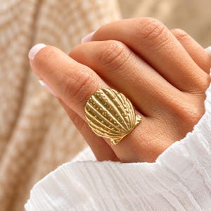 May include: A gold-toned ring featuring a large, fan-shaped seashell design. The surface of the shell is textured with vertical ridges and small, raised dots, creating a detailed pattern that covers the entire front of the ring band.