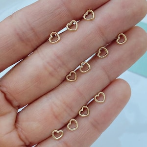 May include: A collection of small, gold-colored heart-shaped stud earrings. The earrings are displayed on a light-colored surface, showcasing their delicate design. The open-heart design is visible in each earring.