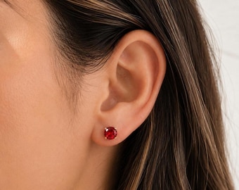 May include: A round, faceted red gemstone stud earring secured in a gold-toned metal setting. The circular stone features a brilliant cut that reflects light, creating a vibrant crimson appearance centred on the earlobe for a classic look.