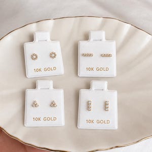 May include: Four pairs of 10K gold stud earrings displayed on white cards. The top left pair features circular designs, the top right pair has horizontal bars, the bottom left pair shows triangular clusters, and the bottom right pair has vertical bars.
