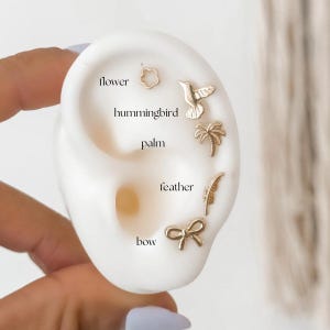 May include: A close-up of a white ear model with gold-colored earrings. The earrings are shaped like a flower, a hummingbird, a palm tree, a feather, and a bow. The earrings are labeled with the words "flower", "hummingbird", "palm", "feather", and "bow".