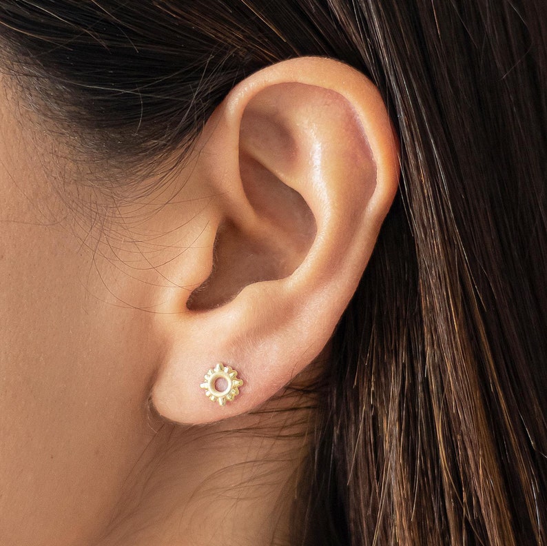 May include: A close-up of a person's ear with a small gold sun earring. The earring has a circular design with a textured, sun-like pattern.