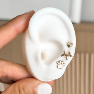 May include: Gold-colored stud earrings displayed on a white ear model. The earrings include a heart, a balloon dog, and a paw print design. The earrings are small and delicate, with a polished finish.