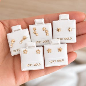 May include: Five pairs of 10kt gold stud earrings displayed on white cards. Designs include shooting stars with clear stones, three-star climbers, simple stars, open-frame stars, and textured starfish. Each card is clearly labeled 10 KT. GOLD.