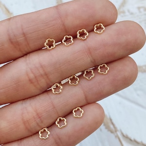 May include: Small, gold-colored flower-shaped earrings. The earrings are arranged in rows on a light-colored surface. The earrings have a delicate, open design, resembling a five-petal flower. The earrings are likely made of metal and are suitable for everyday wear.