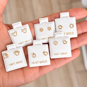 Puede incluir: Seis pares de pendientes de botón de oro de 10kt presentados en tarjetas blancas individuales. Los diseños incluyen siluetas de ratón, estrellas, círculos, flores y corazones. Cada tarjeta blanca tiene el texto 10KT. GOLD en letras doradas.