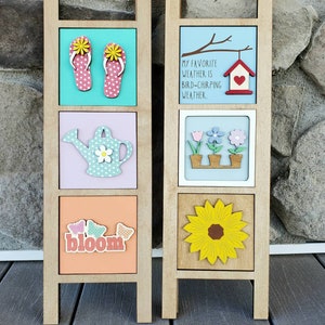 May include: Two wooden ladder-style signs with three square panels each. The left sign has a pair of pink flip flops, a blue watering can, and the word "bloom" with butterflies. The right sign has a blue panel with the text "My favorite weather is bird-chirping weather." and a red birdhouse, a panel with three potted flowers, and a yellow sunflower.