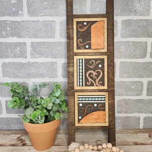 May include: A wooden wall decor with three square panels featuring intricate geometric and floral designs in shades of brown, orange, blue, and white. The panels are attached to a dark brown wooden frame.