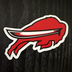 May include: A red and white Buffalo Bills logo patch. The logo features a red buffalo head with a black outline and a white stripe. The patch has a white border and is set against a black background.