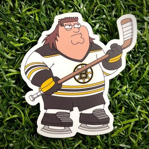 Op de afbeelding: Een sticker met een stripfiguur in een Boston Bruins hockey-shirt, met een hockeystick. Het shirt is wit met zwarte en gouden accenten en het Bruins-logo is zichtbaar. Het personage heeft bruin haar en draagt een bril.