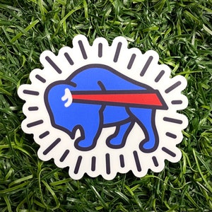 May include: A white sticker with a blue buffalo silhouette and a red stripe across its back. The buffalo is surrounded by a white circle with black rays emanating from it.