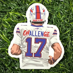 May include: A white football jersey with blue and red accents. The jersey has the number "17" on the back and the words "RISE TO THE CHALLENGE" printed on the back. The jersey is worn by a football player wearing a blue and red helmet.