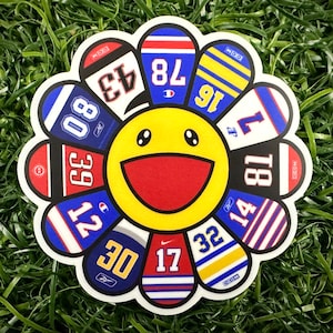 Könnte beinhalten: Ein farbenfroher, blumenförmiger Aufkleber mit einem lächelnden Gesicht in der Mitte. Die Blütenblätter der Blume sind mit verschiedenen Sportteam-Logos und Nummern verziert, darunter 80, 39, 12, 30, 17, 32, 14, 81, 16, 78, 43 und 17.
