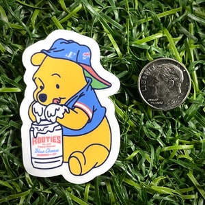 May include: A yellow cartoon bear wearing a blue and green baseball cap and a blue jersey is holding a white can with the text "Rootie's Famous Gourmet Blue Cheese Dressing and Dip".