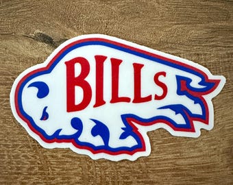 Puede incluir: Una pegatina troquelada que muestra la silueta de un bisonte blanco con un contorno azul y rojo. La palabra BILLS está impresa en letras mayúsculas rojas grandes y negritas en el centro de la forma del bisonte, con un diseño estilizado de cola y cabeza.
