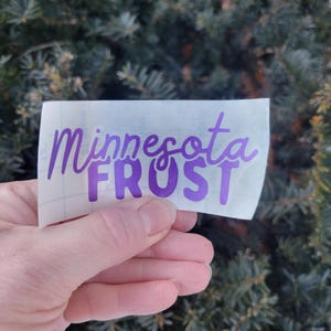 May include: A purple vinyl sticker with the words "Minnesota FROST" in a handwritten font. The sticker is held in front of a background of green evergreen branches.