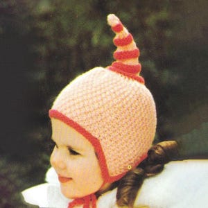 May include: A light pink knitted hat with a red trim and a whimsical, spiralling horn. The hat is designed with a textured stitch and a red tie. The hat is a charming accessory for children.