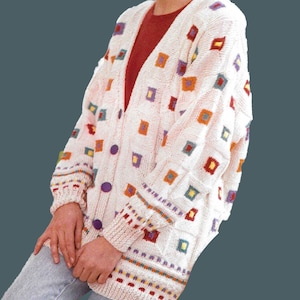 Puede incluir: Un cárdigan blanco con un patrón geométrico colorido. El cárdigan tiene cuello en V y botones en la parte delantera. Las mangas son largas y tienen un patrón de rayas coloridas en los puños.