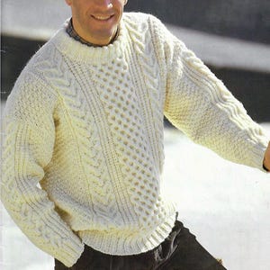 May include: A cream-coloured, cable-knit jumper with a crew neck. The jumper features intricate patterns and textures, with a diamond-like design on the front. The sleeves and body have different cable patterns. The person is wearing dark corduroy trousers.