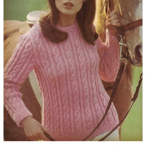 以下が含まれることがあります： ピンクのケーブルニットセーターを着た女性が茶色の馬の横に立っています。彼女は右手に馬の綱を持っています。