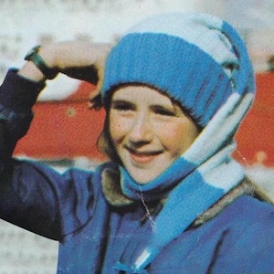 May include: A person wearing a blue knit hat with a light blue stripe and matching scarf. The person is also wearing a blue jacket with a fur-lined collar. A watch is visible on the person's wrist.